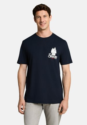 Mann trägt ein schwarzes T-Shirt mit kleinem weißen Snoopy-Hundehäuschen und Text auf der linken Brustseite, kombiniert mit hellgrauer Hose.
