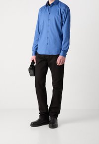 Camicia button-up azzurro chiaro con maniche lunghe, pantaloni neri e scarpe nere. Tiene una piccola borsa nera con tracolla.