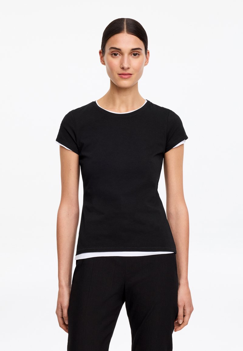 Femme debout, visage tourné vers l'avant, portant un t-shirt noir cintré à manches courtes avec un liseré blanc aux manches et à l’ourlet sur fond blanc.
