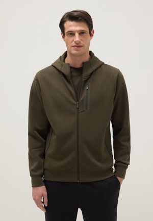 Man met een olijfgroene hoodie met ritssluiting, borstzakken en zijzakken, staand tegen een effen lichte achtergrond.