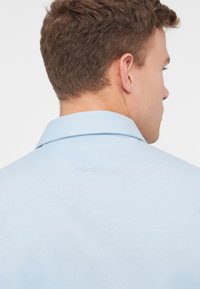 Gaiši zila polo kreka zīmols ar apkakli, kura virsma ir gluda un ar smalkām izšuvuma detaļām. Dizains ir vienkāršs, bez redzamām rakstām.