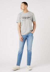 Grey cotton t-shirt featuring the printed logo "Wrangler," paired with light blue denim jeans and white canvas sneakers.
