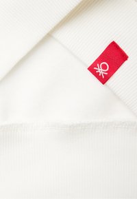 Weiße Baumwollstoff mit weicher Textur, versehen mit einem roten Etikett mit weißem Logo, am Ausschnitt geschichtet mit gerippten Details.