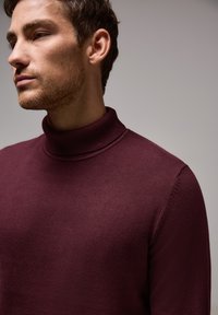 Burgundy rullhalströja i mjukt stickat tyg. Har en ribbad krage och ärmslut, med en slät yta och figursydd design.