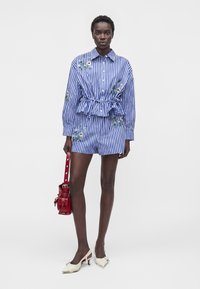 Woman wearing blue and white vertical striped shirt and shorts with floral embroidery, holding a red bag, and white low-heeled shoes.
