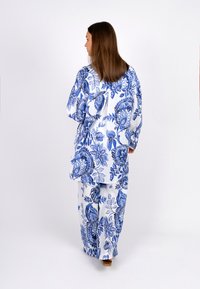 Weißes Stoffoutfit mit aufwendigen blauen Blumenmustern, locker geschnitten, mit langen Ärmeln und weiten Hosenbeinen. Bemerkenswertes Schleifendetail auf der Rückseite.