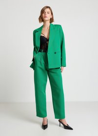 Costume vert sur mesure avec un blazer à un bouton et un pantalon à jambes larges, associé à un haut en dentelle noire et des escarpins noirs à bouts pointus.