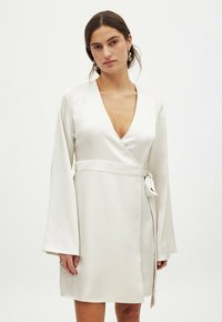 Robe en satin blanc avec un décolleté en V profond, des manches évasées longues et un nœud sur le côté. Texture lisse et design à hauteur du genou.
