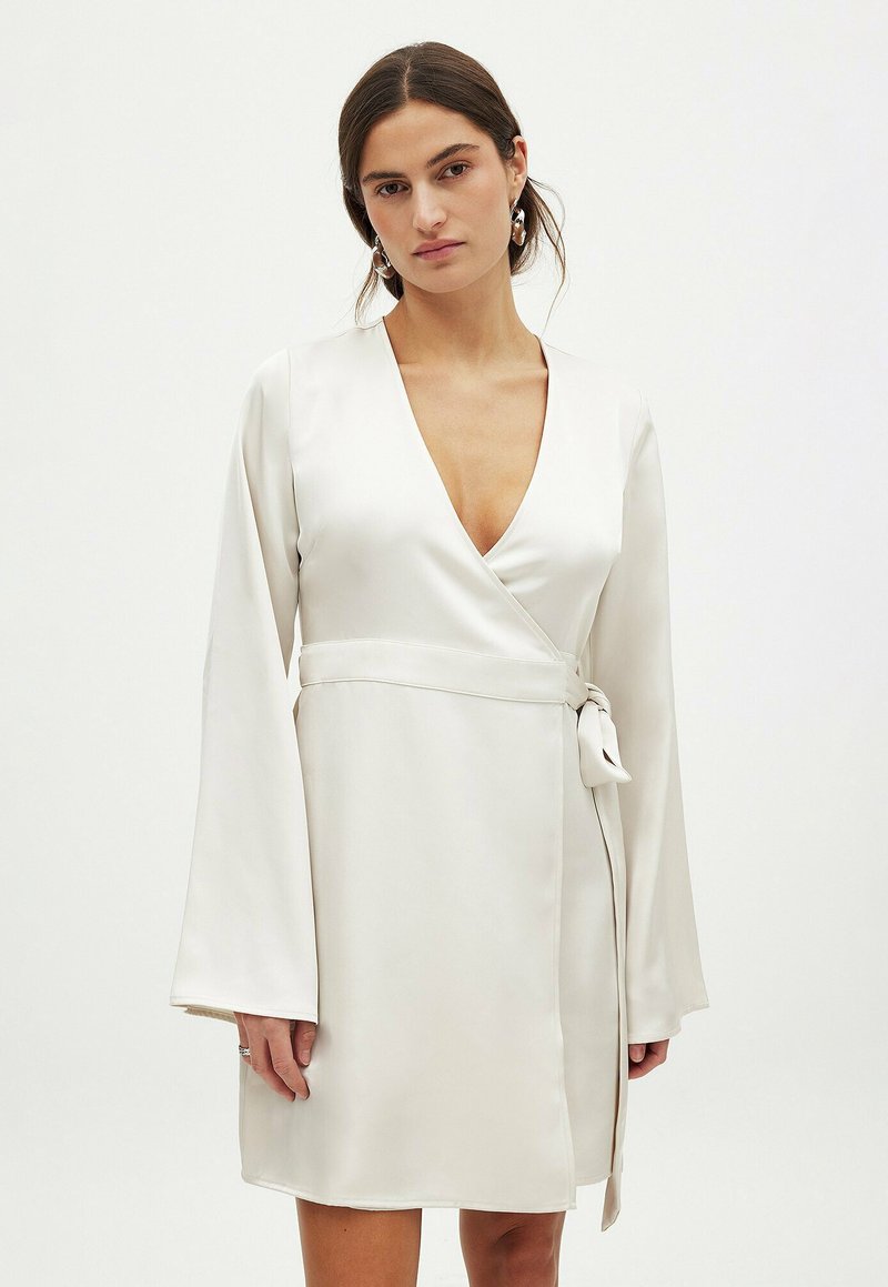 Robe en satin blanc avec un décolleté en V profond, des manches évasées longues et un nœud sur le côté. Texture lisse et design à hauteur du genou.