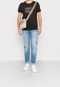 Hombre que lleva una camiseta negra de Tommy Hilfiger, jeans azul claro doblados, zapatillas blancas y un bolso cruzado beige.