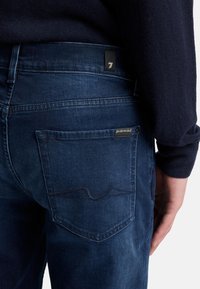 Jeans blu scuro in denim, con una tasca posteriore caratterizzata da un dettaglio a onde cucito e un piccolo etichetta in tessuto sul cinturino.