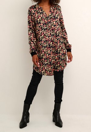 Femme portant une longue tunique à motif floral avec des poignets noirs, un pantalon noir ajusté et des bottines noires à talons, debout devant un fond uni.