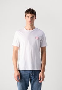 Hvit bomull t-skjorte med rund hals og korte ermer, med en liten rød rektangulær logo på brystet. Kombinert med blå jeans.