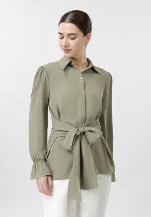 Femeie purtând o bluză de culoare verde măsliniu deschis, cu nasturi, mâneci bufante și o fundă mare legată în talie, asortată cu pantaloni albi.