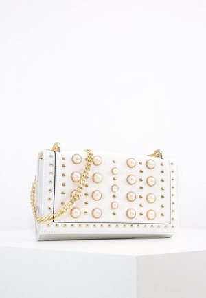 Bolso blanco texturizado con tachuelas doradas y grandes adornos de perlas, con una correa de cadena dorada, colocado sobre una superficie blanca.