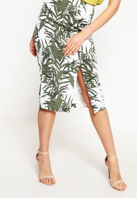 Vit midikjol med ett grönt lövmönster, med en sidoklyvning och en figurnära design. Bärs med beige högklackade sandaler.