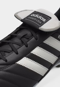 Μαύρο ποδοσφαιρικό παπούτσι Adidas Kaiser Liga με λευκές ρίγες και ετικέτα στη γλώσσα με το λογότυπο Adidas και το κείμενο Kaiser Liga.