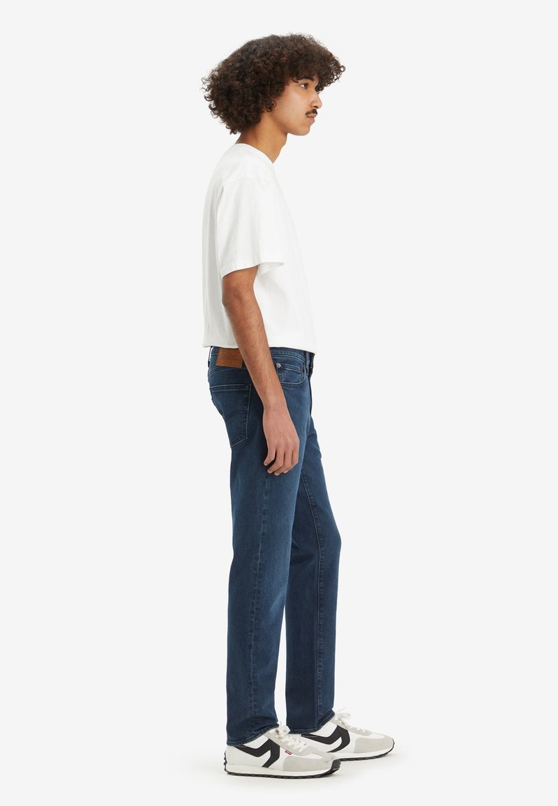 Jeans bleu foncé avec une coupe droite et une étiquette en cuir marron, associés à un t-shirt blanc à manches courtes et des baskets noir et blanc.