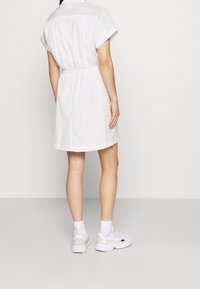 Kobieta ubrana w białą sukienkę z krótkim rękawem i dopasowanym w pasie, białe skarpetki do kostek oraz białe sneakersy na grubej podeszwie, stojąca na jednolitym tle.