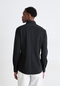 Camisa negra de manga larga con tejido texturizado, cuello estándar y puños con botones. Presenta una ligera curvatura en el dobladillo y detalles en el yugo trasero.