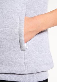 Sweatshirt gris avec un tissu doux et texturé, une poche kangourou et un ourlet côtelé. Le design comprend un cordon de serrage dissimulé pour les ajustements.