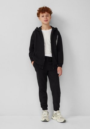 Zwarte ritssluiting hoodie over een wit T-shirt, gecombineerd met zwarte joggers en beige sneakers. De outfit heeft een eenvoudig ontwerp zonder zichtbare patronen.
