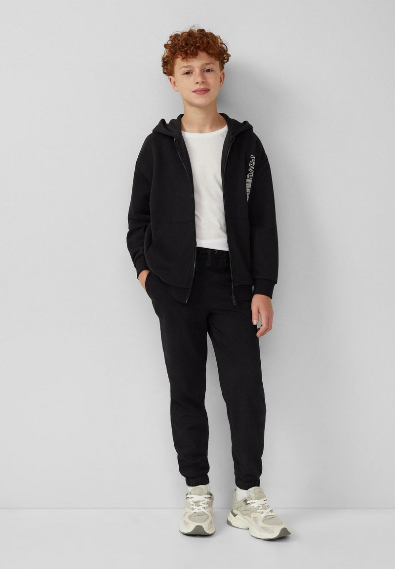 Zwarte ritssluiting hoodie over een wit T-shirt, gecombineerd met zwarte joggers en beige sneakers. De outfit heeft een eenvoudig ontwerp zonder zichtbare patronen.