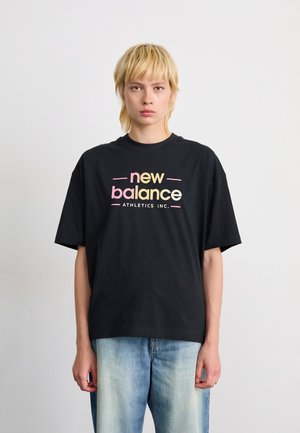 Personne avec les cheveux blonds courts portant un t-shirt noir de New Balance Athletics Inc. et un jean bleu clair devant un fond blanc uni.