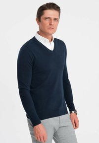 Maglione blu navy a scollo a V con maniche lunghe, texture a maglia, polsini e orlo a coste, abbinato a una camicia bianca con colletto e pantaloni grigi.