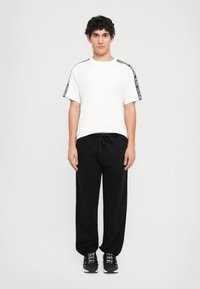 Jovem em pé, vestindo uma t-shirt branca com riscas pretas na altura dos ombros, calças pretas com cordão e ténis pretos com texto branco.