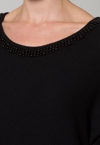 Maglione nero lavorato a maglia con scollatura ampia, caratterizzato da un accento di perline lungo il colletto. Il tessuto appare morbido con una trama testurizzata.