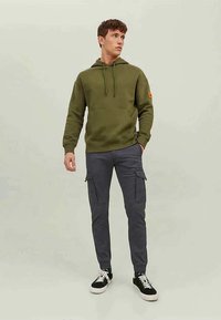 Hoodie verde-oliva com cordões e bolso frontal, combinado com calças cargo cinza-escuras com bolsos laterais. Ténis preto e branco.