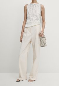 Biały dzianinowy top na ramiączkach z misternym wzorem, w połączeniu z luźnymi kremowymi spodniami i teksturowaną szaro-brązową torbą.