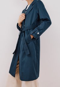 Marineblauer Trenchcoat mit taillierter Gürtelschließe, seitlichen Taschen und knopfbaren Manschetten, aus glattem, wasserabweisendem Stoff gefertigt.