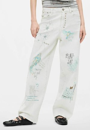 Persona con vaqueros blancos y sueltos decorados con bocetos, frases escritas a mano y pájaros coloridos; zapatos planos negros con correas y hebillas.