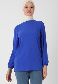 Maglietta blu a maniche lunghe con una vestibilità rilassata, scollo rotondo e polsini elasticizzati, abbinata a jeans azzurri. Tessuto morbido e leggero.