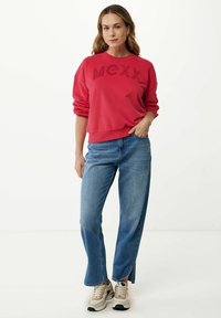 Sudadera roja con el logo "MCXX", escote redondo y dobladillo acanalado. Combinada con jeans de pierna ancha azules con aberturas laterales y zapatillas beige.