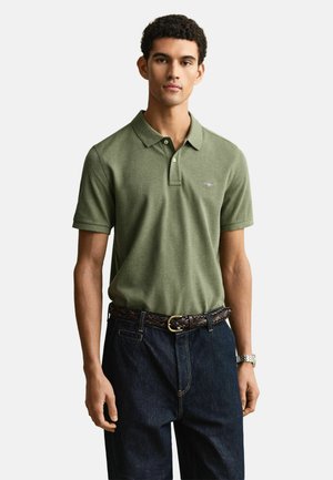 Jonge man met een olijfgroen poloshirt, donkerblauwe spijkerbroek met een gevlochten bruine riem en een zilveren polshorloge, staand tegen een witte achtergrond.
