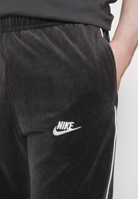Černé velurové joggery s elastickým pasem, bočními kapsami a vyšitým logem Nike v bílé barvě, s bílým lemováním podél švů.