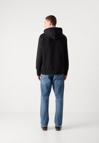 Felpa nera con cappuccio ampio, polsini a costine, indossata con jeans diritti blu chiaro e scarpe marroni, vista da dietro.