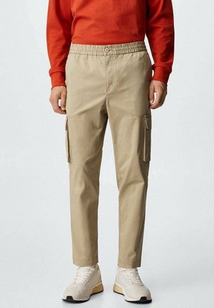Mand iført beige cargobukser med sidelommer, hvide sneakers og en rød langærmet skjorte, stående mod en ensfarvet baggrund.