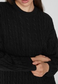 Schwarzer Kabelstrickpullover mit rundem Halsausschnitt und gerippten Bündchen, der ein strukturiertes Muster aufweist und eng am Körper sitzt.