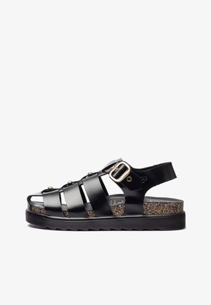 Sandal de tiras negras con un acabado brillante, que presenta un acento de hebilla, plantilla de corcho y suela texturizada negra. Diseño de punta abierta.