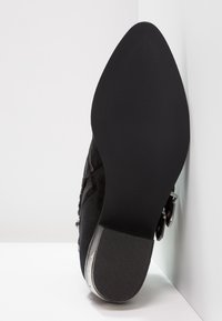 Scarpa in suede nero con suola in gomma liscia, punta affusolata e un accento metallico vicino alla caviglia. Presenta una chiusura con zip sul lato.