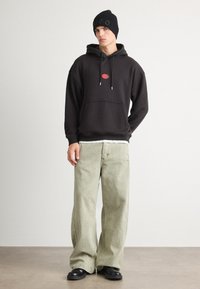 Felpa nera con cappuccio e logo rosso, pantaloni larghi verde chiaro e berretto nero. Texture elegante con vestibilità comoda. Footwear neri completano il look.