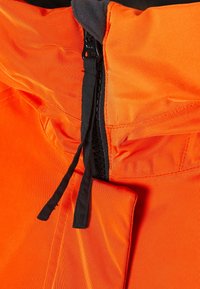Gros plan sur la fermeture éclair avant d'une veste orange partiellement ouverte avec des tirettes noires et des détails de couture visibles.
