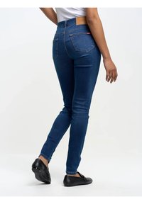 Sieviete, kas valkā pieguļošas zilas džinsu bikses un melnas loafer kurpes, stāvot ar vienu pēdu nedaudz paceltu uz vienkārša baltā fona.