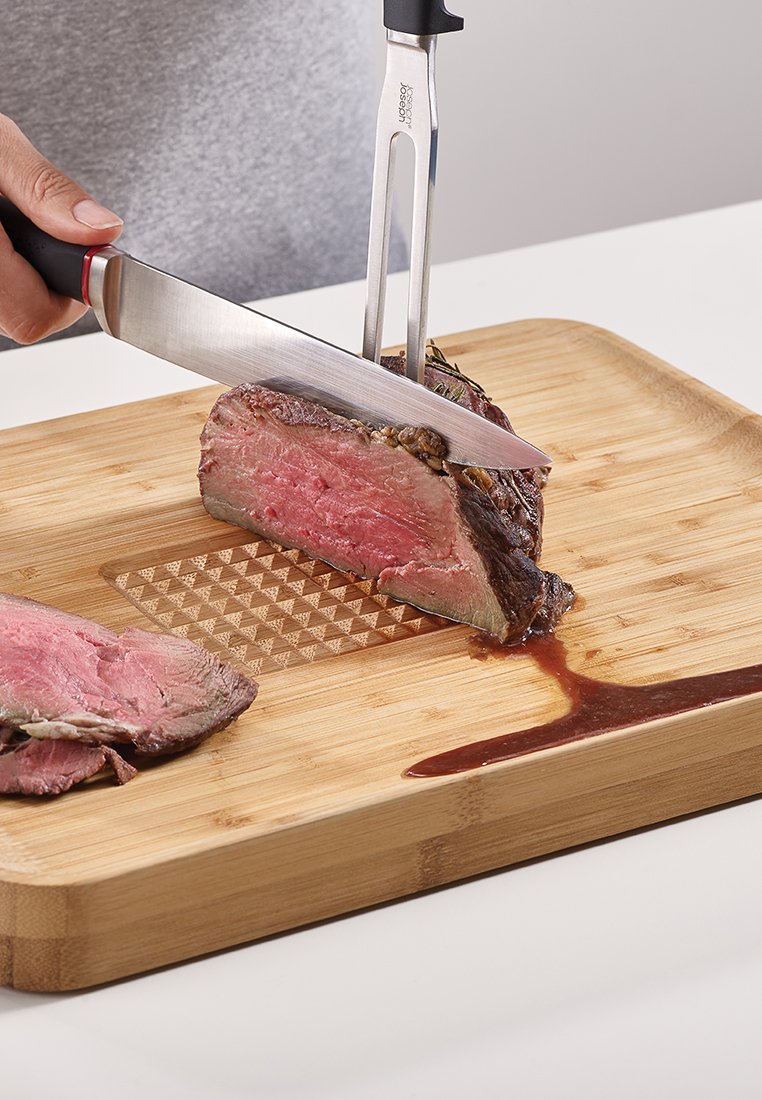 Ein Schneidemesser und eine Gabel, die ein medium-rare Steak auf einem Bambusschneidebrett schneiden, wobei das rosa Fleisch und der braune Saft auf der glatten Oberfläche zu sehen sind.