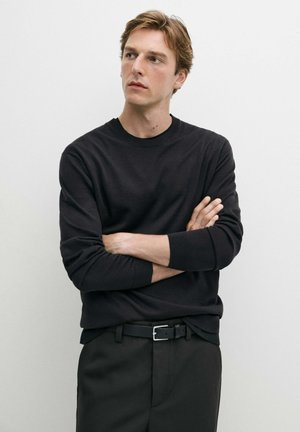 Mand med lyst brunt hår iført sort sweater og bukser står med armene over kors og kigger til siden mod en ensfarvet hvid baggrund.