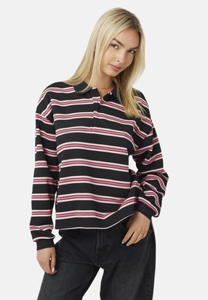 Giovane donna con lunghi capelli biondi che indossa una polo a maniche lunghe a righe nere, rosa e bianche e pantaloni neri, in piedi su sfondo bianco.